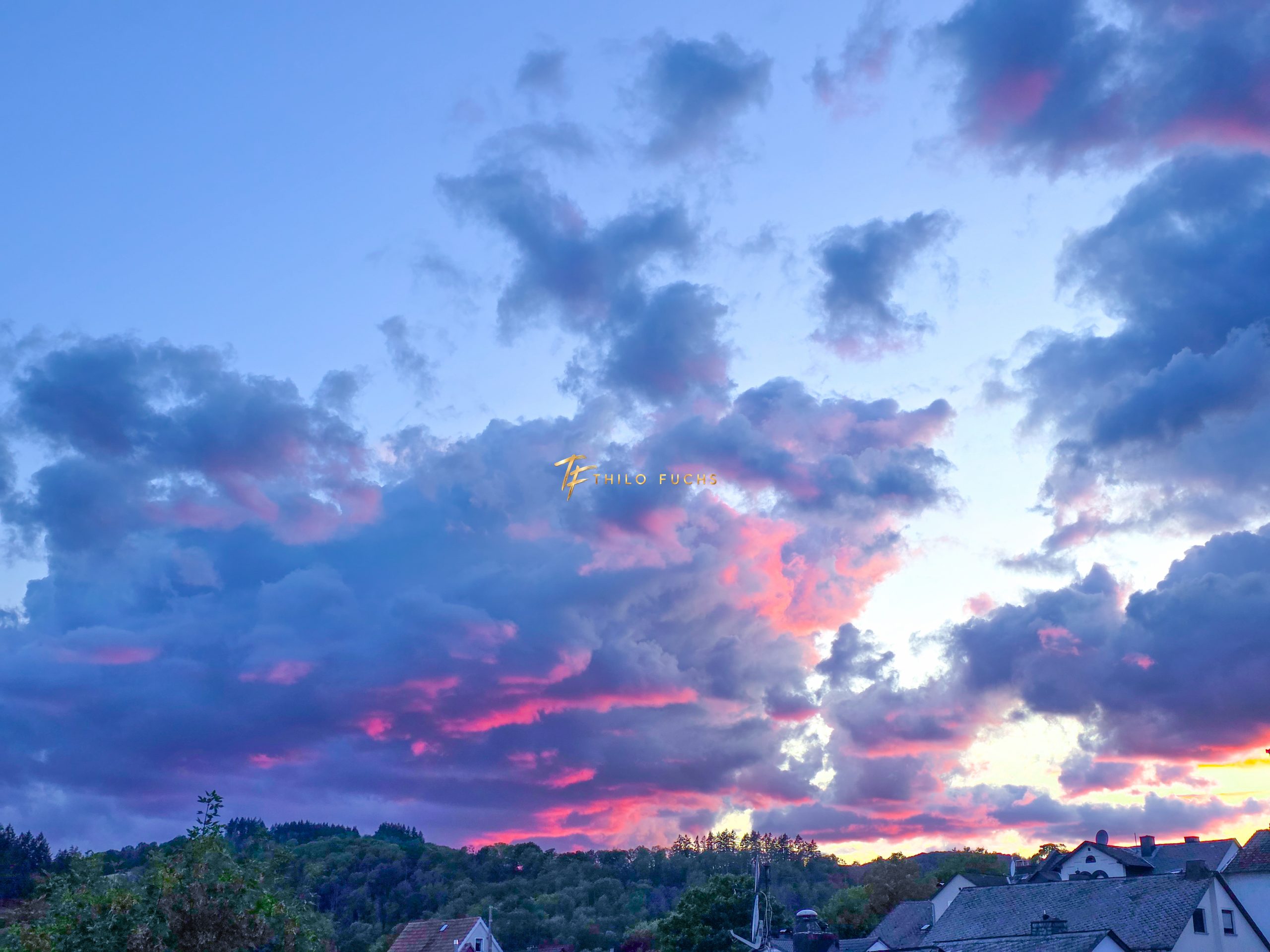 Zwischen Dunkelheit und Licht: Ein Abendhimmel in fünf Momenten Blauer Abendhimmel mit rosa beleuchteten Wolken über Häusern und bewaldetem Horizont.
