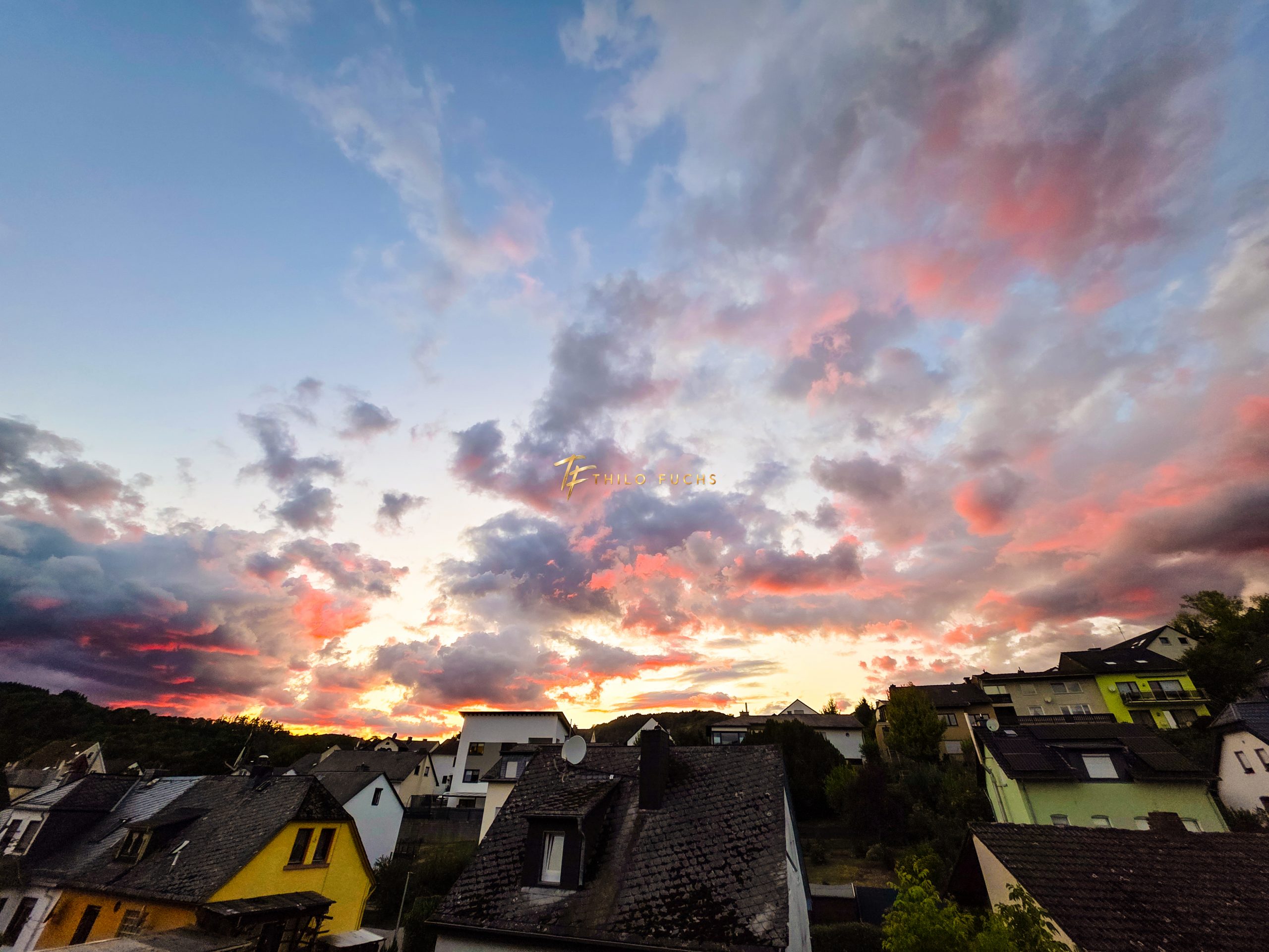 Zwischen Dunkelheit und Licht: Ein Abendhimmel in fünf Momenten Weiter Blick über Hausdächer auf einen Sonnenuntergang mit rosa, roten und goldenen Wolken.